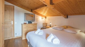 A bedroom with a wooden ceiling, a bed with white linens, a wooden dresser, a chair, and a mirror on the wall.