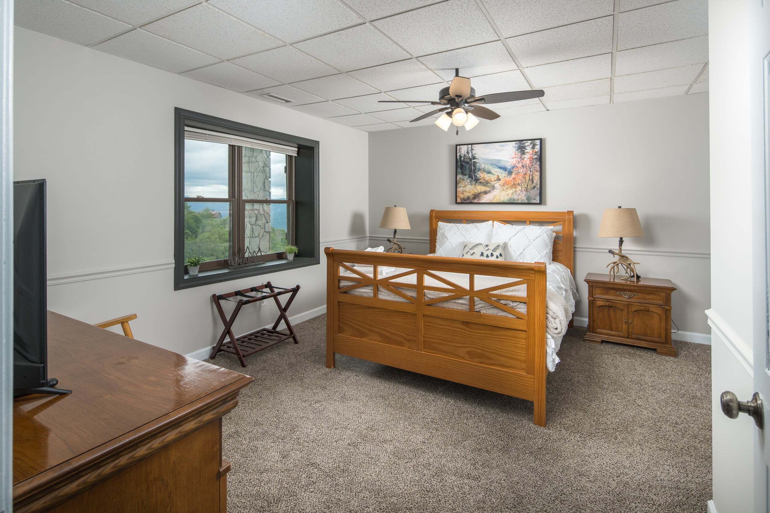 Quarto com cama de madeira, ventoinha no teto, janela com vista, escrivaninha e mesa de cabeceira.