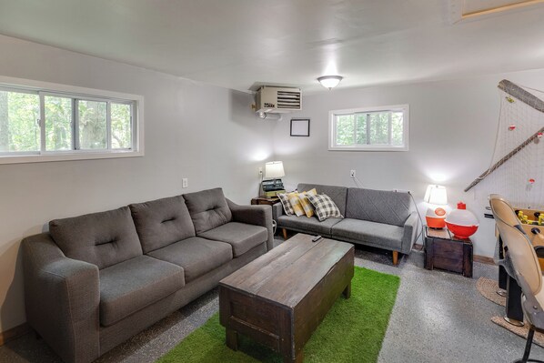 A living room with two grey sofas, a wooden coffee table, and a green rug. There are two windows, a ceiling fan, and a lamp on a side table.