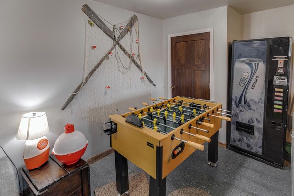 A room with a foosball table, a refrigerator, and a lamp on a wooden table.