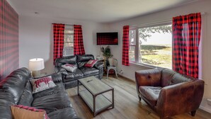 A living room with a leather sofa, a coffee table, and a TV. There are plaid curtains on the windows.