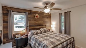 A bedroom with a bed, a ceiling fan, a nightstand with a lamp, a dresser, and a window with curtains.