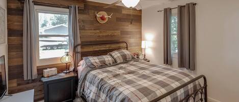 A bedroom with a bed, a ceiling fan, a nightstand with a lamp, a dresser, and a window with curtains.