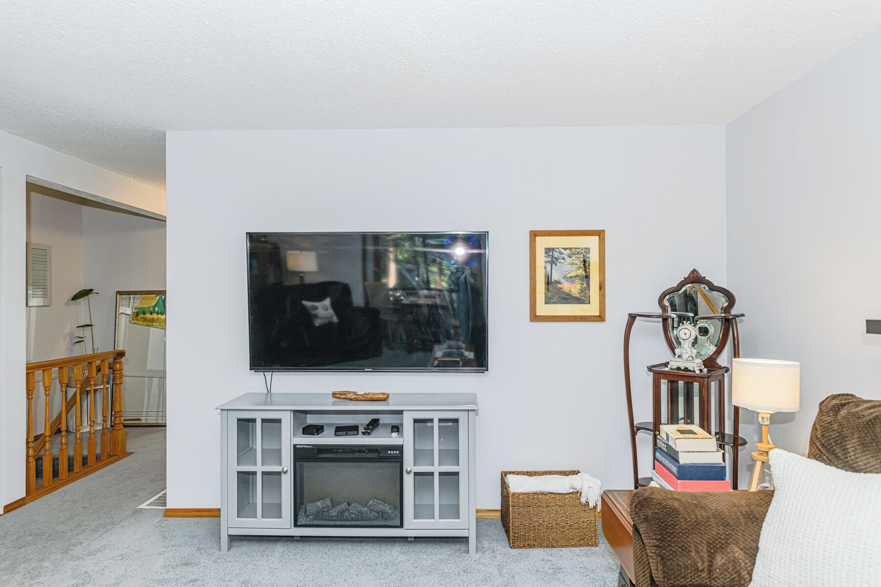 Uma sala de estar com televisão de tela plana na parede, um móvel branco com lareira, uma cesta de vime e uma poltrona marrom.
