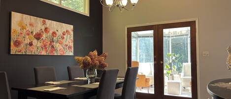 A dining area with a dark wooden table, grey chairs, a vase with dried flowers, and a large floral painting on the wall.