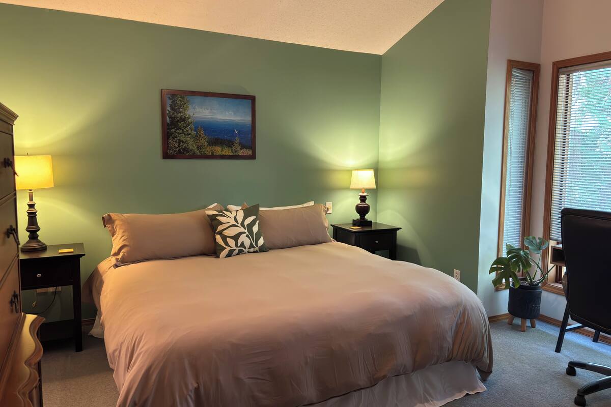 A bedroom with a bed, two bedside tables with lamps, a nightstand, a potted plant, and a window with blinds.