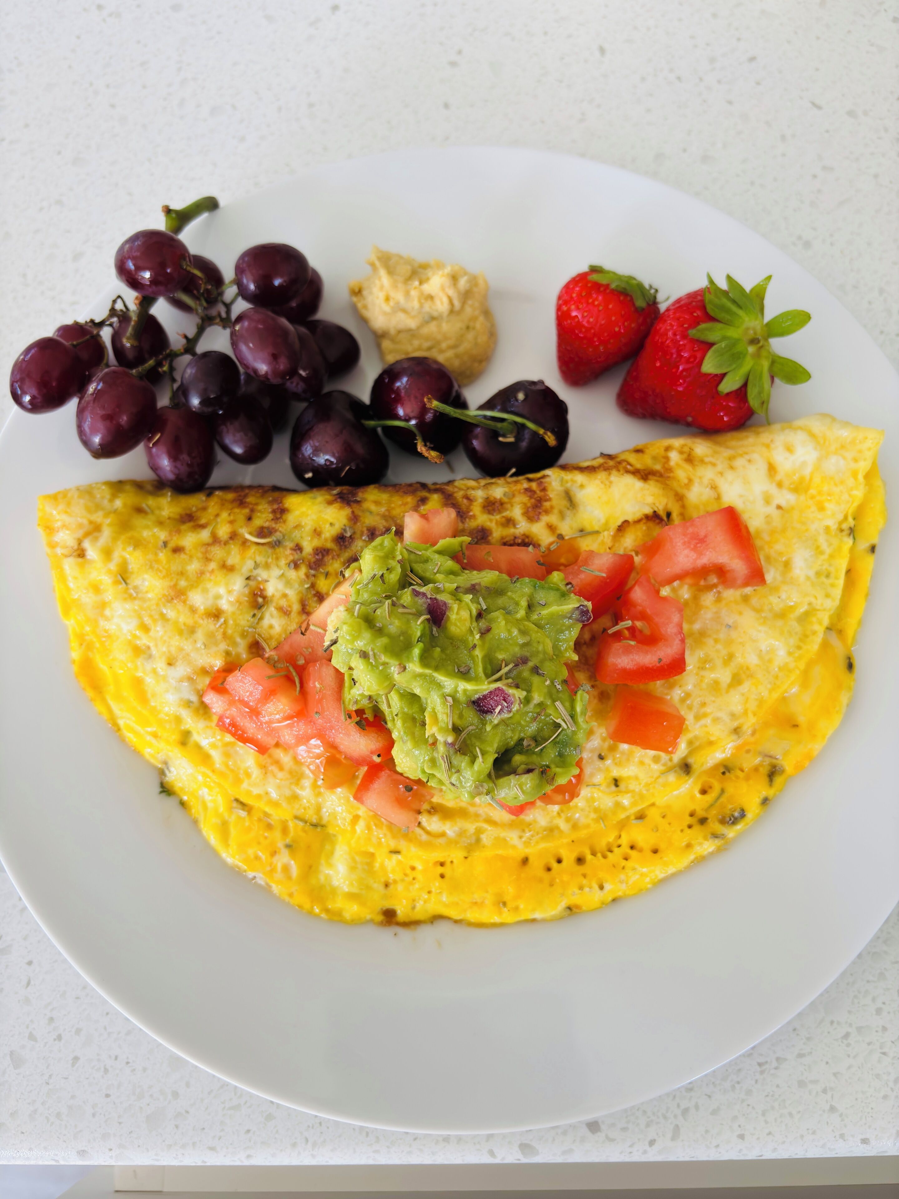 A plate with an omelette, grapes, cherries, strawberries, and a dollop of guacamole.