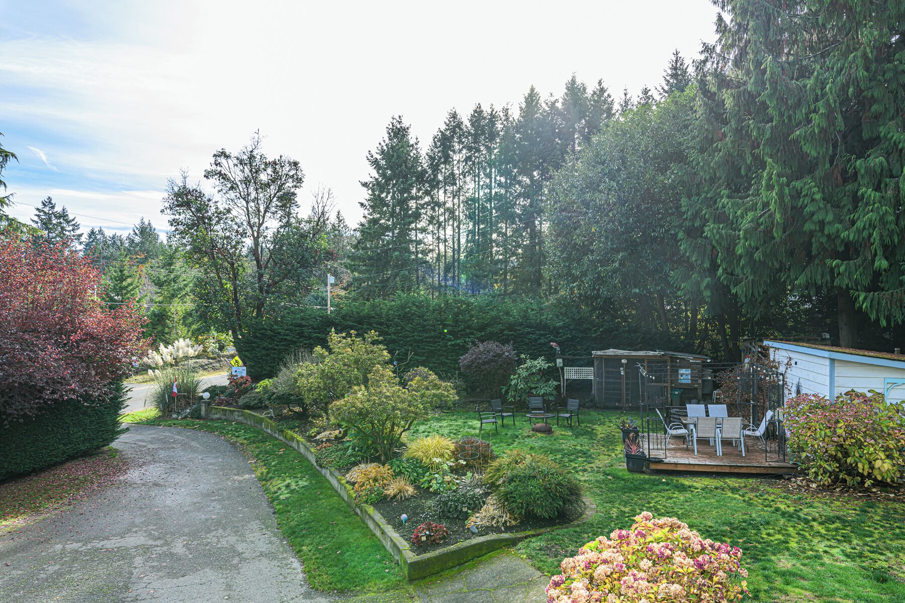 A residential garden with a paved driveway, a patio area with outdoor furniture, and a variety of plants and trees.
