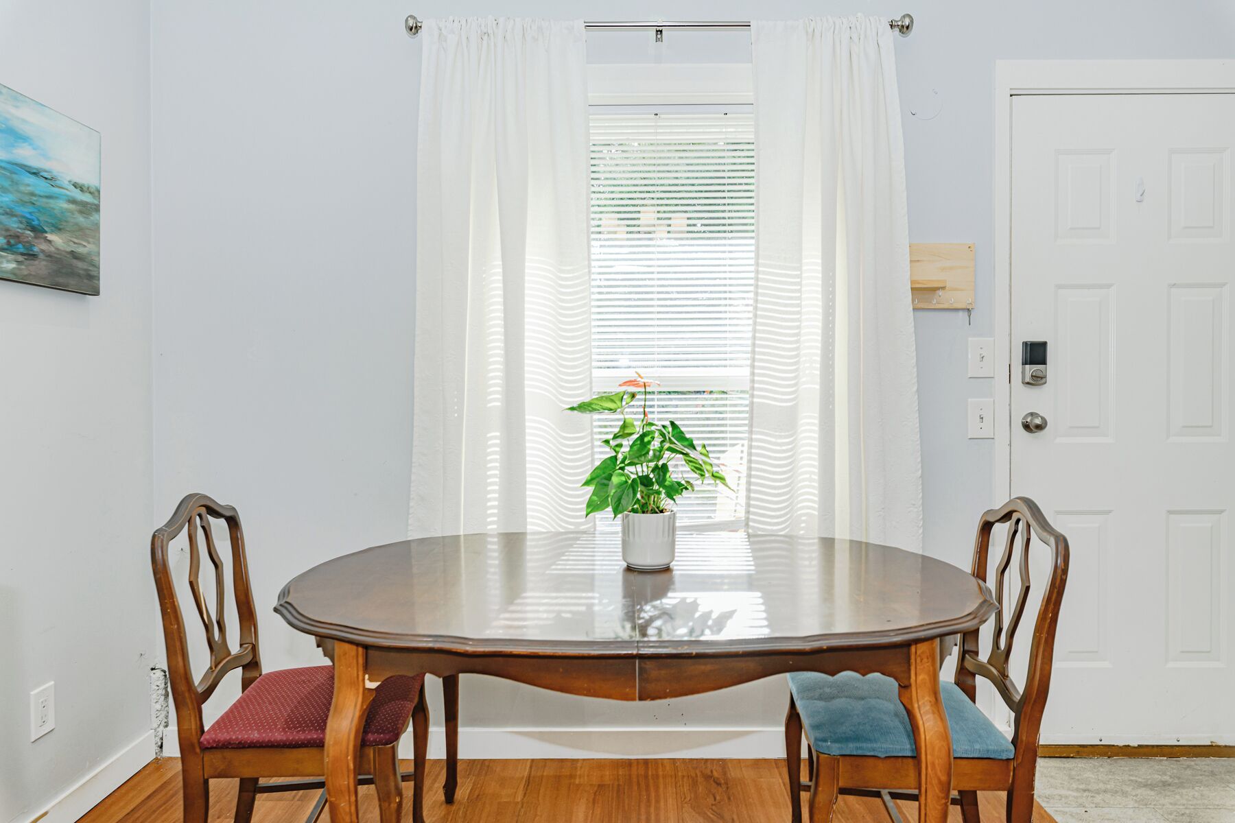 A dining area with a round wooden table, two chairs, a potted plant, and a window with blinds.