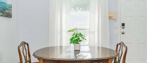 A dining area with a round wooden table, two chairs, a potted plant, and a window with blinds.