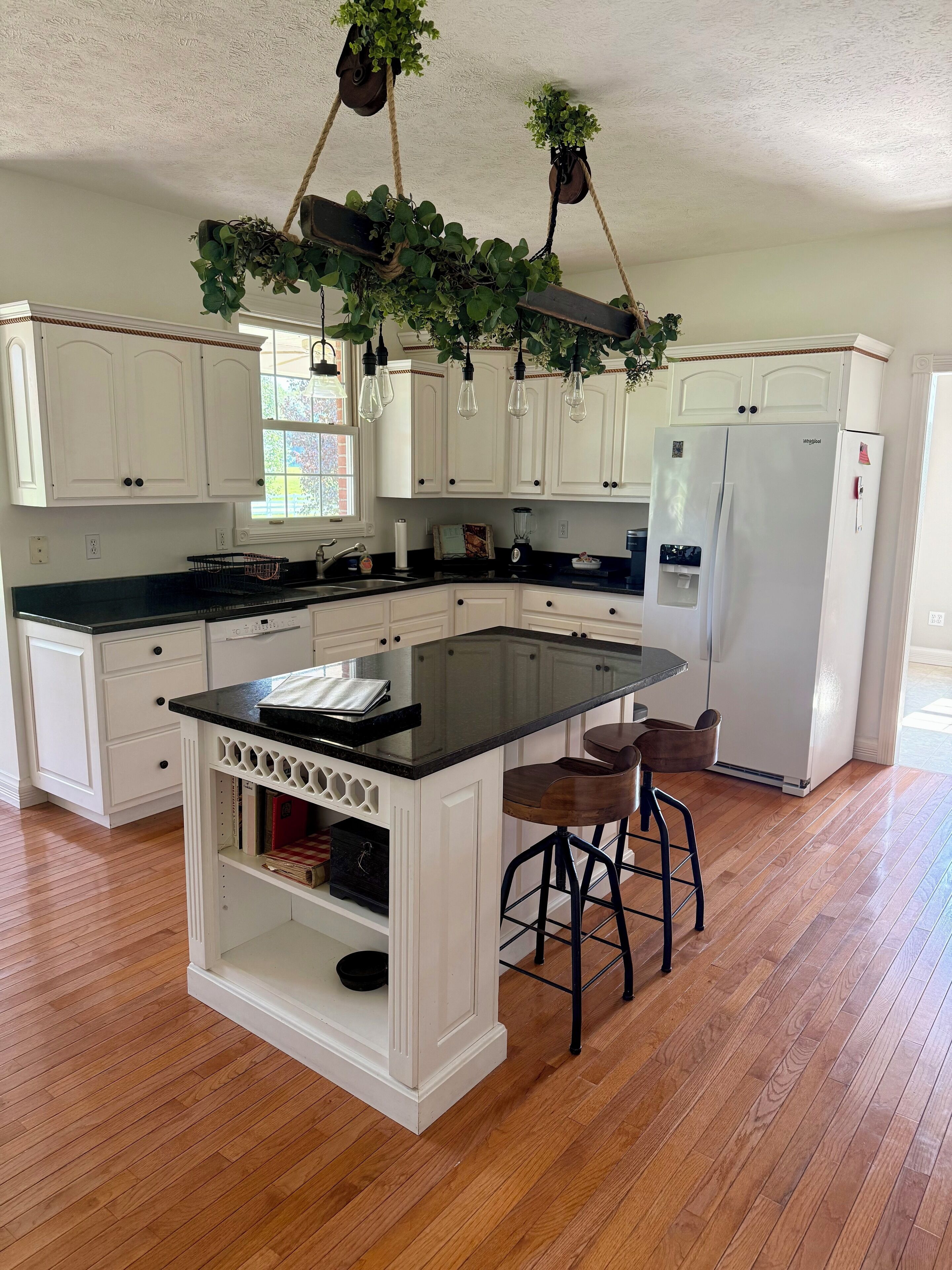 A kitchen with white cabinets, a black countertop, a central island with stools, and hanging plants.