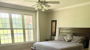 Une chambre à coucher comprenant un lit, une table de chevet, un ventilateur de plafond et une fenêtre donnant sur un paysage verdoyant.
