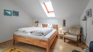 Un dormitorio con una cama de madera, una mesita de noche pequeña con una lámpara, una silla y un tragaluz sobre la cama.