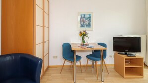 Una habitación pequeña y moderna con piso de madera, una mesa de comedor con dos sillas, un televisor sobre un mueble de madera y un ventanal grande.