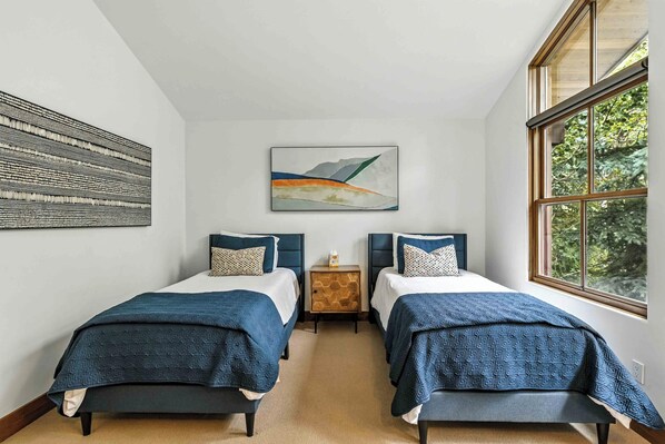 A room with two beds, a small wooden nightstand, a painting, and a window with a view of trees.