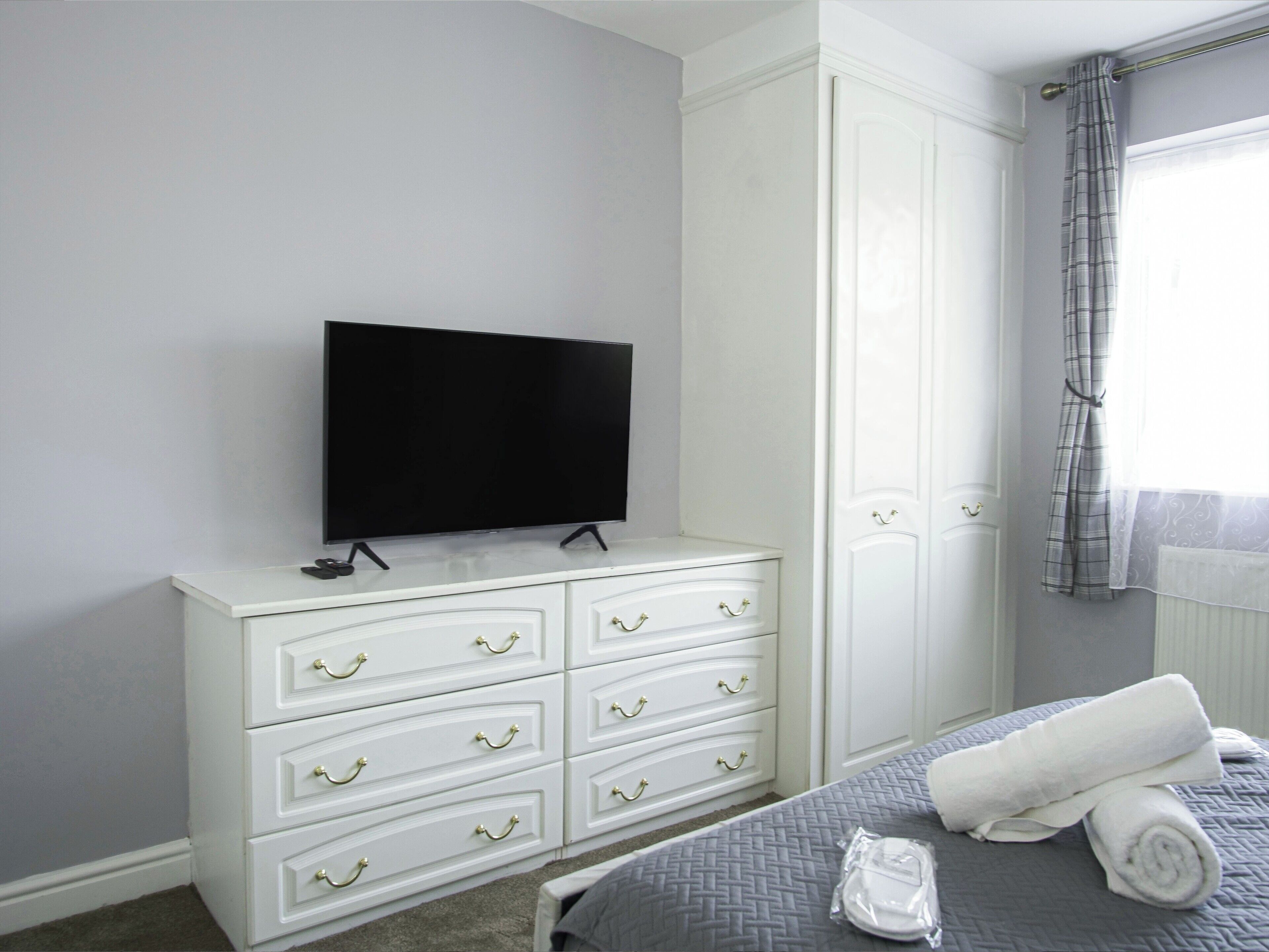 A bedroom with a flat-screen TV on a white dresser, a bed with a grey quilt, and a window with a plaid curtain.