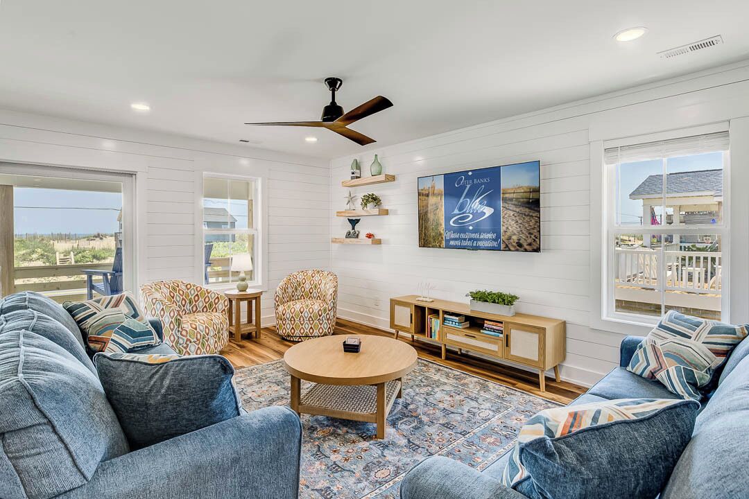 A spacious living room with a large sofa set, a coffee table, and a TV mounted on the wall. There is a ceiling fan, a bookshelf, and a window with a view of the outdoors.