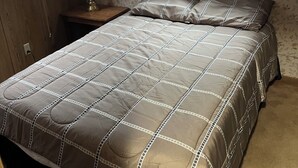 Een slaapkamer met een bed, nachtkastje, lamp en raam met jaloezieën.