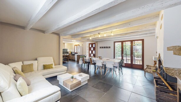 Un vaste espace de vie comprenant un canapé blanc, une table basse et un coin repas avec une table et des chaises. Au fond, on aperçoit une cuisine équipée d’un téléviseur et de placards.