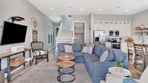 A modern living room with a sectional sofa, two round coffee tables, a flat-screen TV, and a staircase leading to an upper level. - 2 Mins to Beach | Pool View Retreat | Fully Loaded (Gulf Shores)