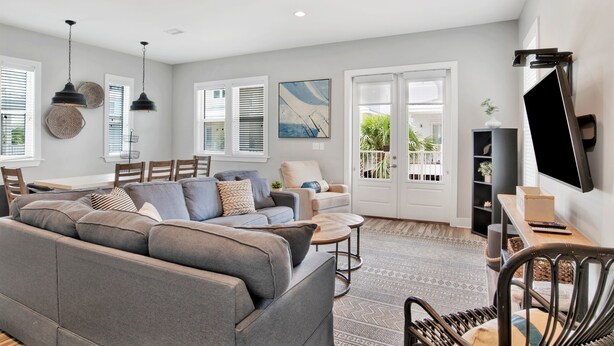 A modern living room with a grey sectional sofa, a coffee table, and a TV on a stand. There are two pendant lights, a painting on the wall, and a door leading to a balcony. - 2 Mins to Beach | Pool View Retreat | Fully Loaded (Gulf Shores)