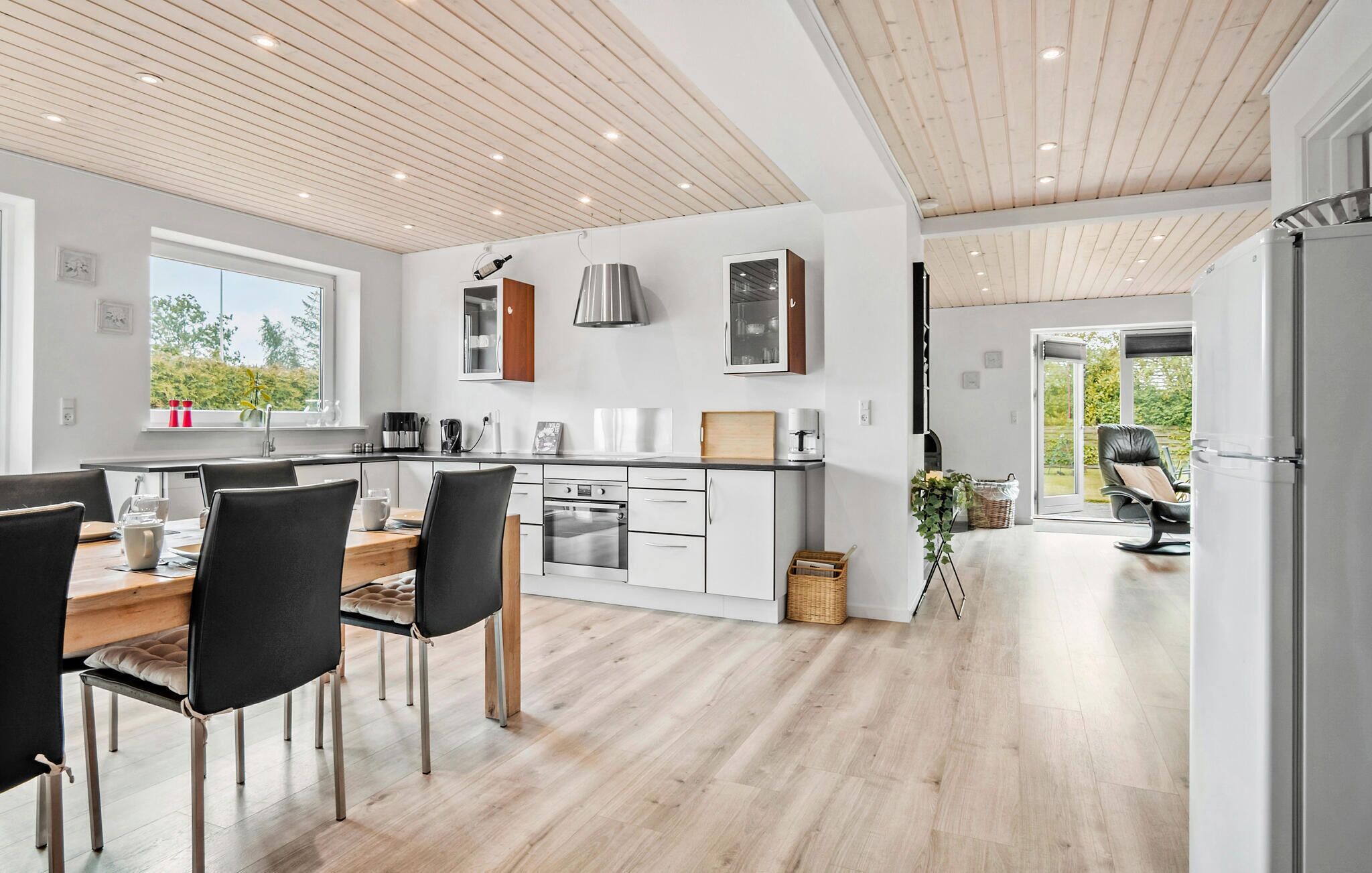 Une cuisine moderne avec des meubles blancs, un coin repas avec des chaises noires et une vue sur la verdure à l’extérieur.