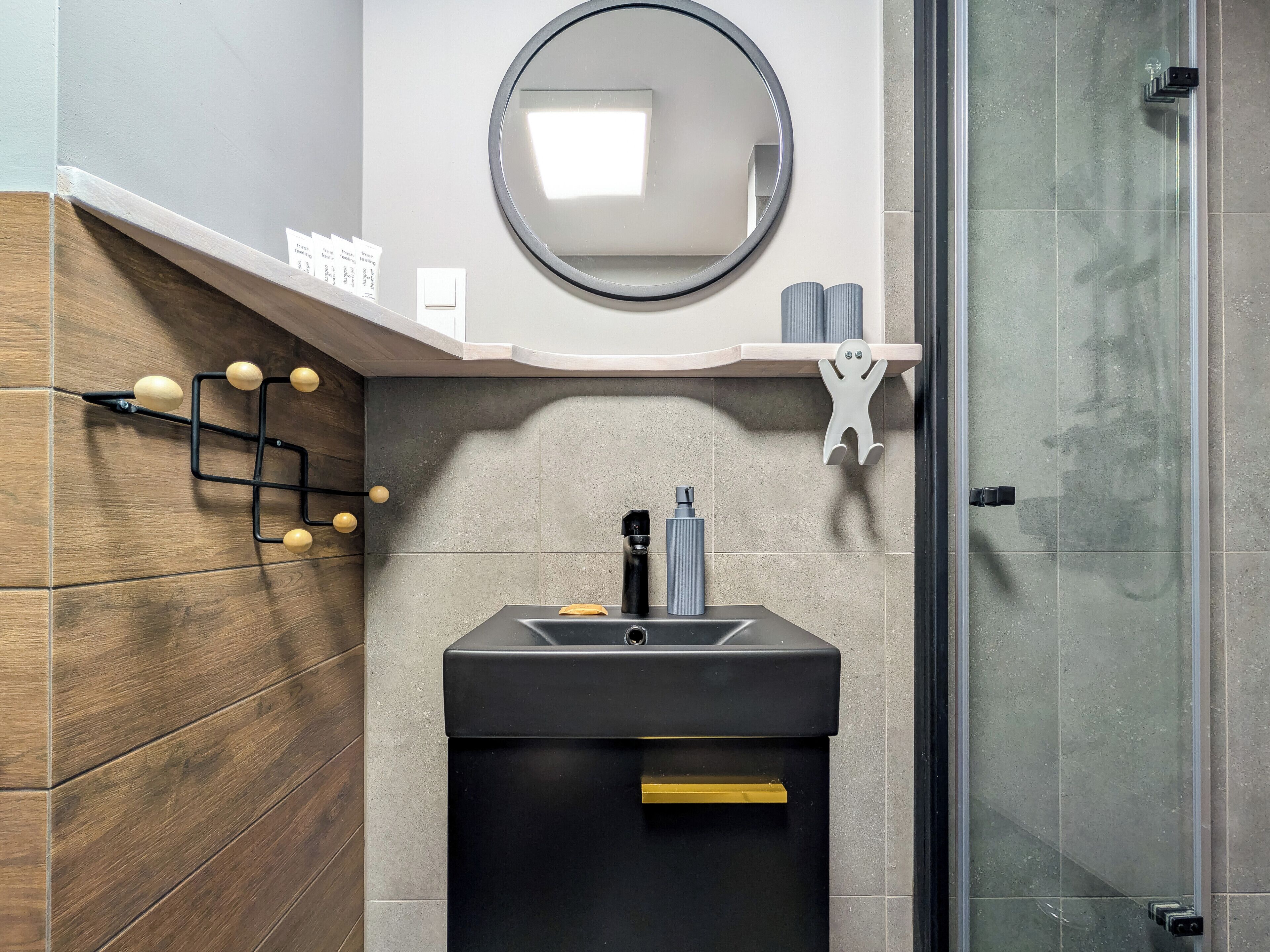 Un baño moderno con pared de madera, espejo redondo, lavamanos negro y cabina de ducha de vidrio.