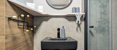 Un baño moderno con pared de madera, espejo redondo, lavamanos negro y cabina de ducha de vidrio.