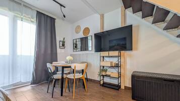 En moderne stue med spiseplads, trægulv, et fladskærms-tv monteret på væggen og en trappe, der fører til en etage ovenpå.