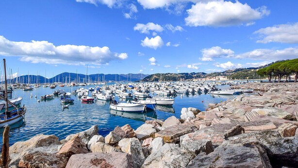 A marina with numerous boats docked, rocky shore in the foreground, and a clear blue sky with scattered clouds. - Pet friendly apartment (Santo Stefano Di Magra)