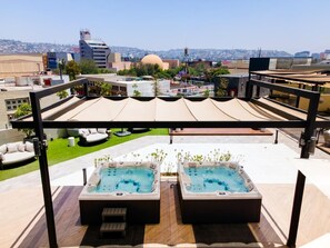A rooftop terrace with two hot tubs, a covered seating area, and a view of the cityscape.