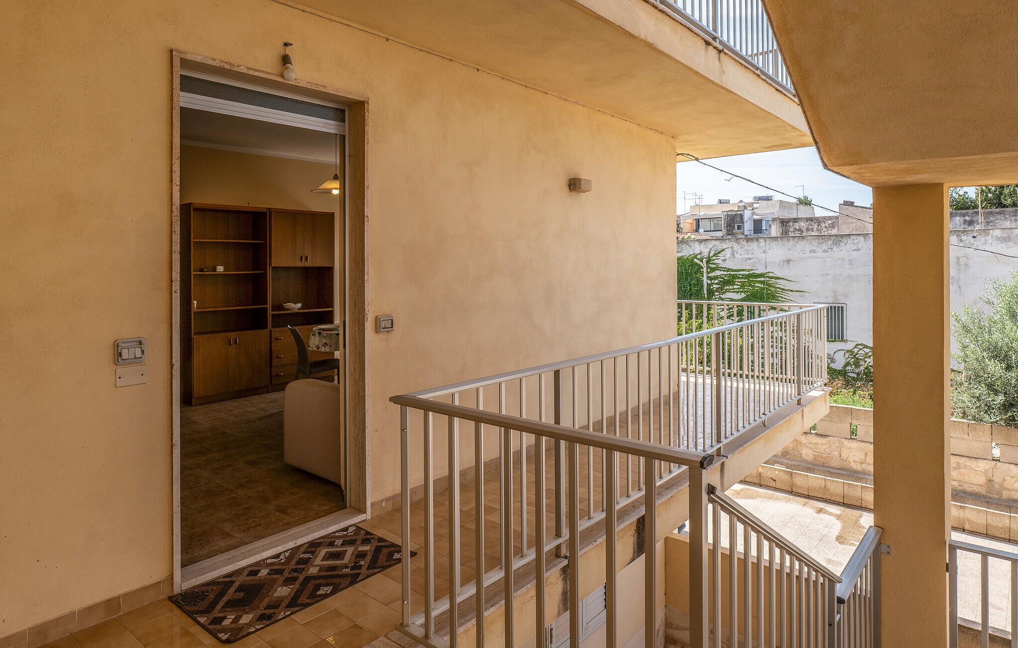 Un balcon d’habitation doté d’une rambarde en métal, une porte donnant sur une pièce intérieure avec un meuble en bois et une chaise, et une vue sur le quartier environnant.