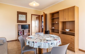 A dining area with a round table set for four, surrounded by chairs. There is a television on a shelf to the left and a wooden cabinet with shelves to the right.