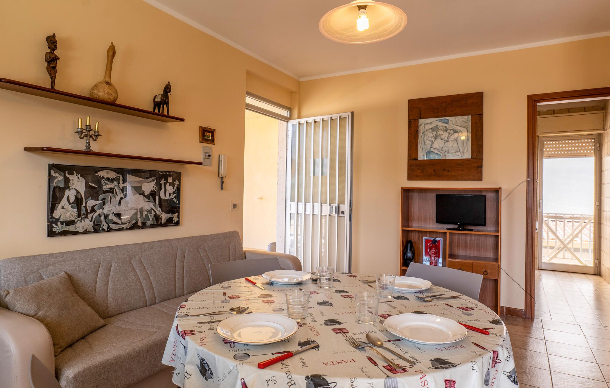 Un coin repas avec une table ronde dressée pour quatre personnes, un canapé, un téléviseur posé sur une étagère et des œuvres d’art accrochées aux murs.