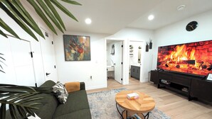 A living room with a sofa, a coffee table, a flat-screen TV displaying a fireplace, and a kitchen in the background. - Luxury Guest Suite in Calgary perfect for a visit to the Canadian Rockies. (Calgary)