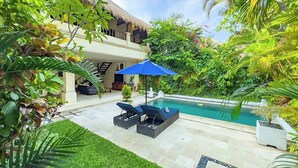 Un área de piscina con sillas de descanso y una sombrilla azul, rodeada de exuberante vegetación y un edificio con techo de paja.