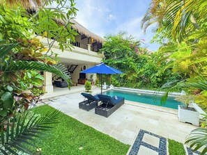 Uma área de piscina com espreguiçadeiras e um guarda-sol azul, cercada por vegetação exuberante e um edifício com telhado de palha.