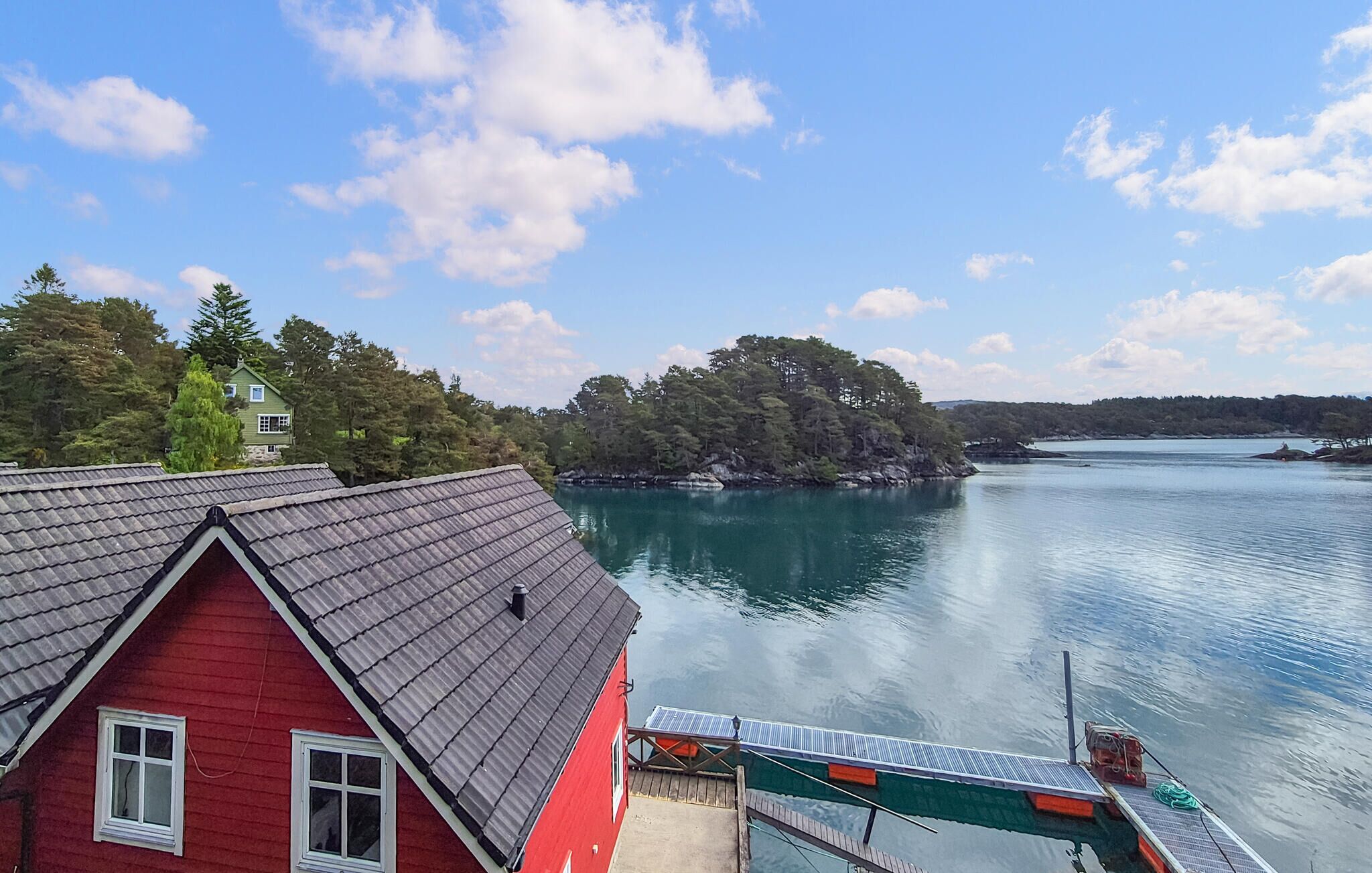 A red house with a sloped roof and a dock extending into a calm lake, surrounded by trees and a clear sky.