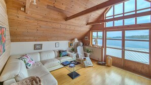 L’intérieur chaleureux d’un chalet avec un plafond en bois, un canapé blanc, une table basse en verre et une grande fenêtre donnant sur l’eau.