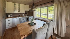 Ein Holzinterieur mit Esstisch, grau gepolsterten Stühlen, einer Küche mit weißen Schränken und einem großen Fenster mit Vorhängen.