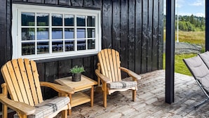 Eine Holzterrasse mit zwei Adirondack-Stühlen und einem kleinen Tisch, ein Fenster mit Seeblick und eine schwarze Außenwand.