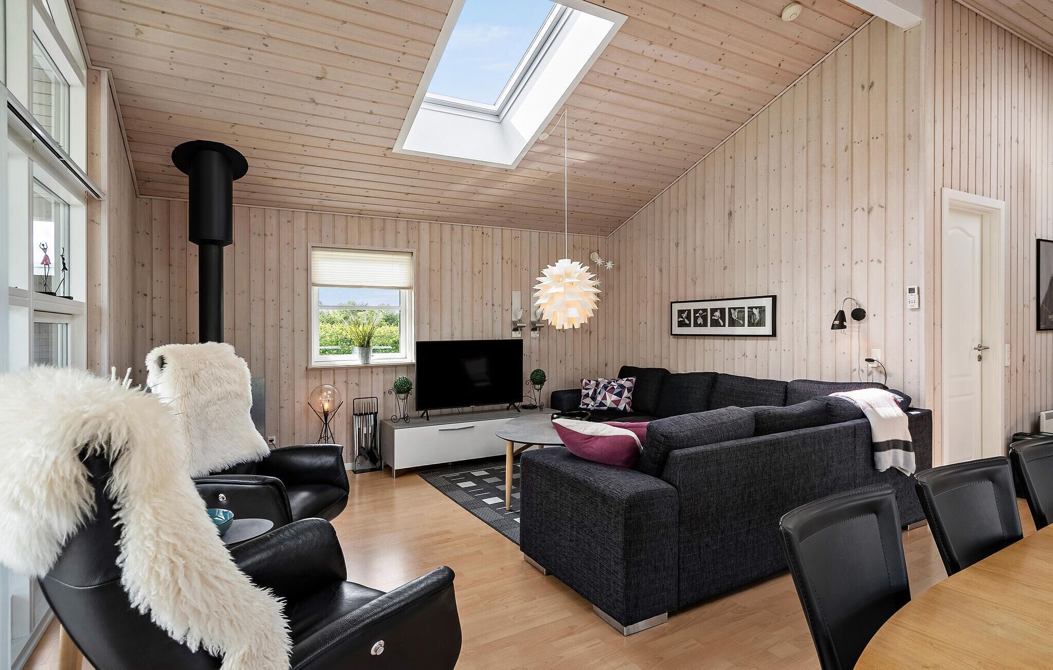 Una sala de estar moderna con un televisor grande, sofás negros, un perro blanco y esponjoso, y un techo de madera con tragaluces.