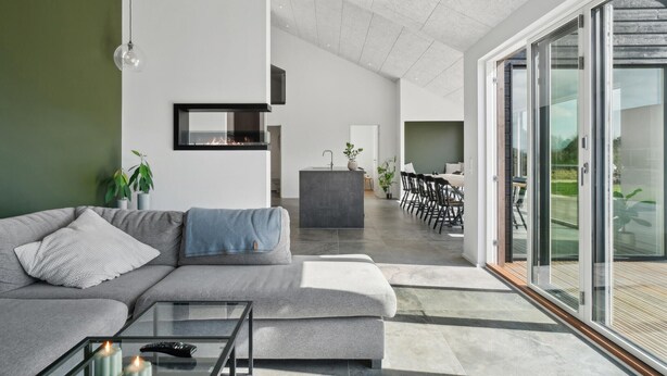 A modern living room with a grey sofa, a glass coffee table, and a fireplace. There is a dining area with a table and chairs visible through a glass door. - 5 bedroom gorgeous home in Skjern (Skjern)