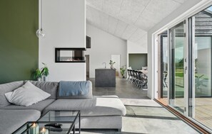 Una sala moderna con un sofá gris, una mesa de centro de vidrio y una chimenea. Se ve un comedor con una mesa y sillas a través de una puerta de vidrio.
