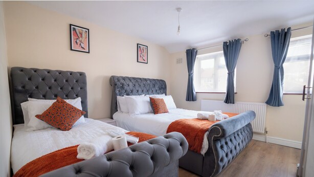 A hotel room with two beds, each with a tufted headboard, white bedding, and orange throw pillows. There are two framed pictures on the wall, a window with blue curtains, and a radiator under the window. - 3-bed House - Sleeps 7, Parking & Views (Belvedere)