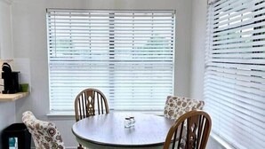 A dining area with a round table, four chairs, a hanging light fixture, and large windows with blinds.