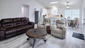 A spacious living room with a brown leather sofa, a round coffee table, and two beige armchairs. A ceiling fan and recessed lighting are visible.
