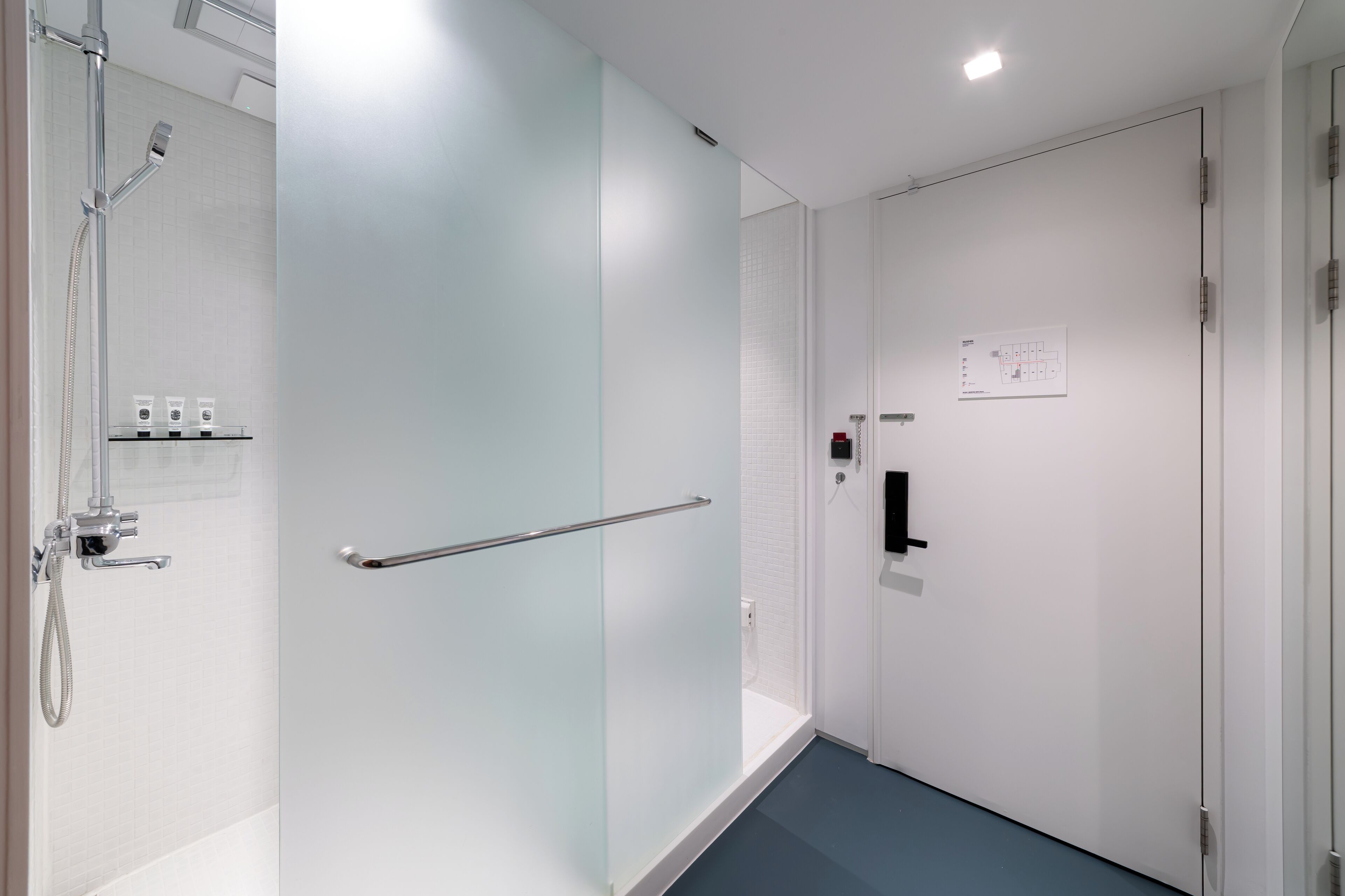 A modern bathroom with a glass shower enclosure, a handrail, and a white door with a control panel.