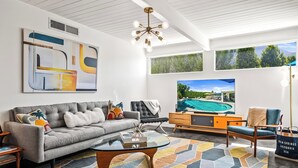 Una sala de estar moderna con un sofá gris, una mesa de centro de vidrio y una gran pantalla de televisión que muestra una escena de piscina.