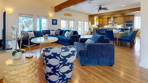 A spacious living room with a high ceiling, wooden beams, a large sofa set, a dining area with blue chairs, and a kitchen in the background.